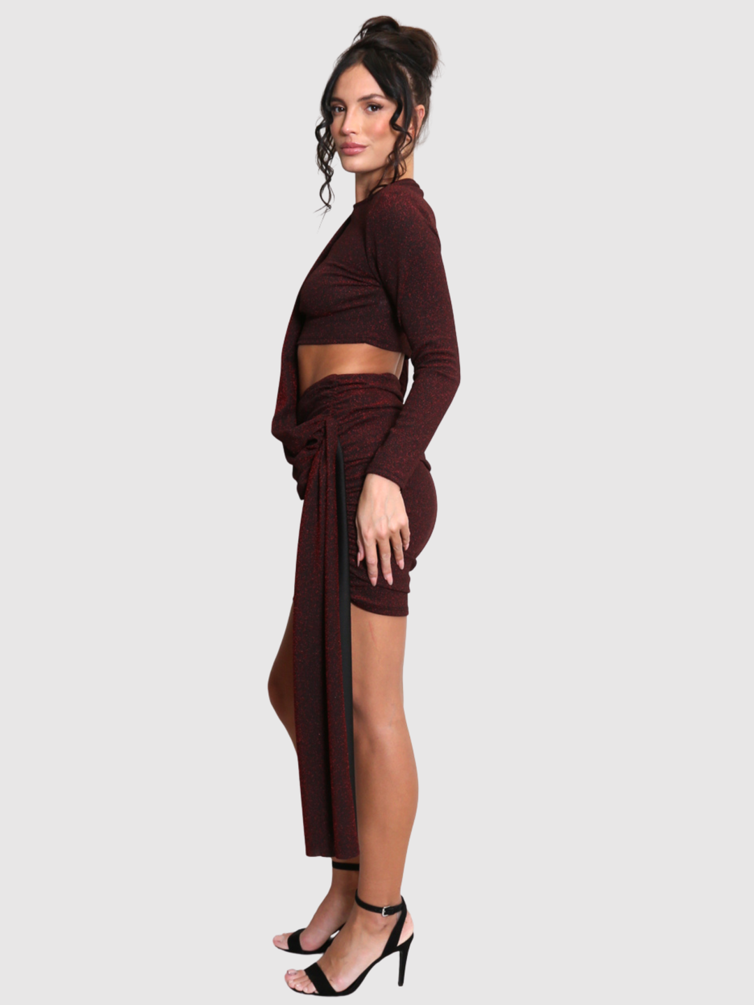 Full length shot. Model stands in front of a plain background. The model wears a cut out lurex burgandy mini dress, with long sleeves and diagnal draped front and rounded neck. The model has tanned skin and black hair that is in a high bun, she stands to the side and turns her head to look into the camera, the draped design can be seen.m 