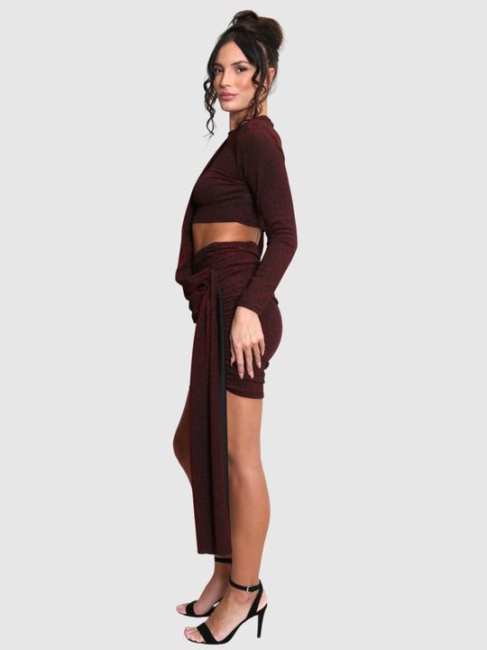 Full length shot. Model stands in front of a plain background. The model wears a cut out lurex burgandy mini dress, with long sleeves and diagnal draped front and rounded neck. The model has tanned skin and black hair that is in a high bun, she stands to the side and turns her head to look into the camera, the draped design can be seen.m 