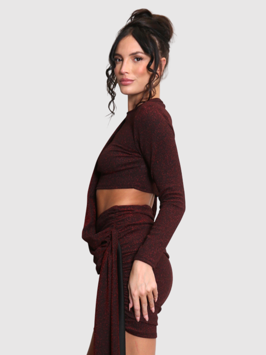 Model stands in front of a plain background. The model wears a cut out lurex burgandy mini dress, with long sleeves and diagnal draped front and rounded neck. The model has tanned skin and black hair that is in a high bun, the model stands to the side and turns her head to look into the camera, the side of the dress can be seen. 