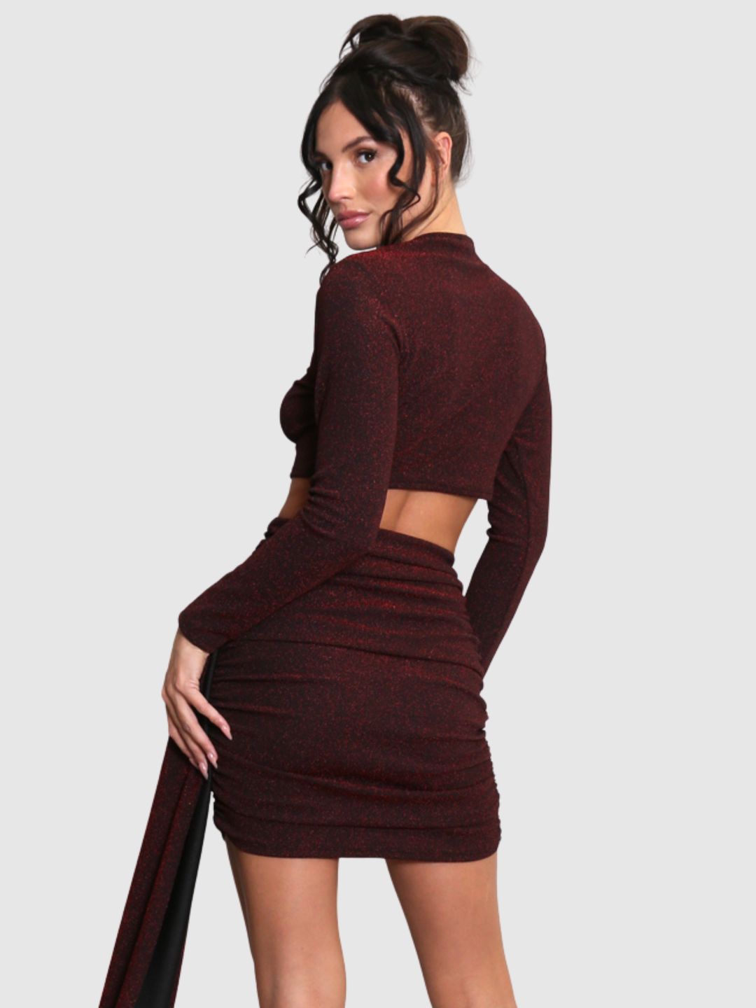Model stands in front of a plain background. The model wears a cut out lurex burgandy mini dress, with long sleeves and diagnal draped front and rounded neck. The model has tanned skin and black hair that is in a high bun, she stands with her back to the camera and turns her head to look into the camera, the back of the  dress can be seen, including the cut out mid drift.