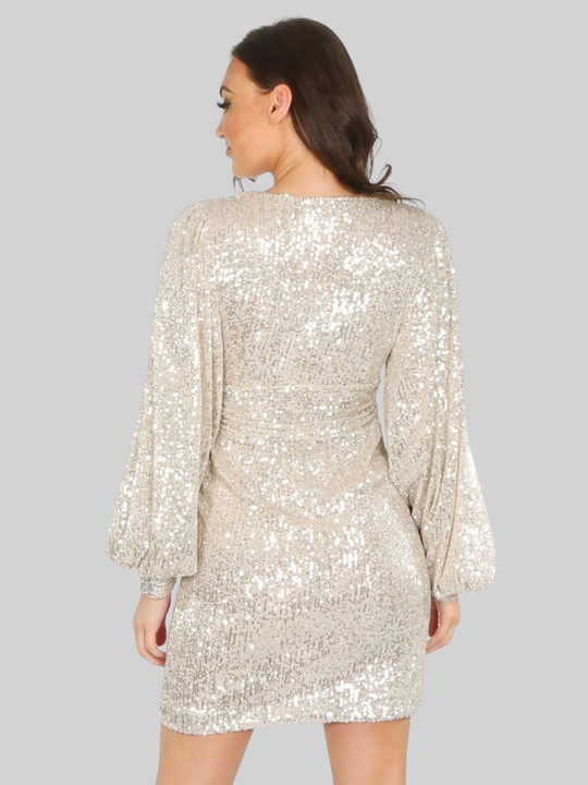 Model stands in front of a plain background. Model wears a champayne sequin mini dress, with v neck front, empire waist amd puffy sleeves. The model has tanned skin and long brown hair, she stands ithher back to the camera, the back of the dress can be seen, demosnstrating the mini dres design.