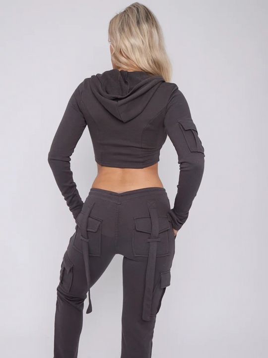 Model wears a cropped top zip up  hoodie in  charcoal grey and matching drawstring joggers with a fitted cargo design. Model has blonde hair,  she stands with her back to the camera, the cargo pants and loop back strings at the back of the joggers can be seen Model places both hands in her pockets. 
