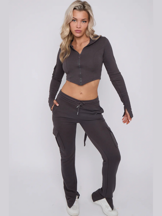 Full length shot. Model wears a cropped top zip up  hoodie in  charcoal grey and matching drawstring joggers with a fitted cargo design. Model has blonde hair, model stands with one hand by her side and the other in her pocket, she looks directly into the camera. Model wears white trainers. 