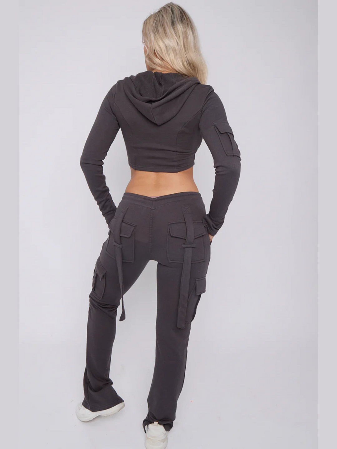 Full length shot. Model wears a cropped top zip up  hoodie in  charcoal grey and matching drawstring joggers with a fitted cargo design. Model has blonde hair,  she stands with her back to the camera, the loop back design of the cargo fitted joggers can be seen. The carfo sleeve of the hooded crop top is also visible. 