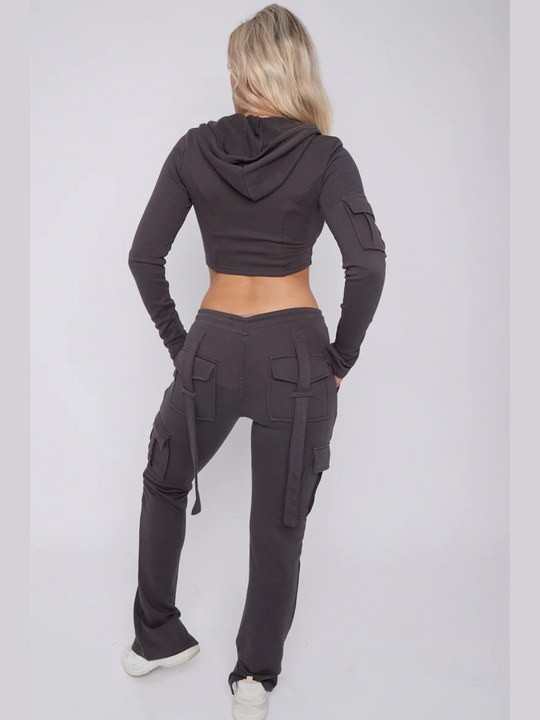 Full length shot. Model wears a cropped top zip up  hoodie in  charcoal grey and matching drawstring joggers with a fitted cargo design. Model has blonde hair,  she stands with her back to the camera, the loop back design of the cargo fitted joggers can be seen. The carfo sleeve of the hooded crop top is also visible. 