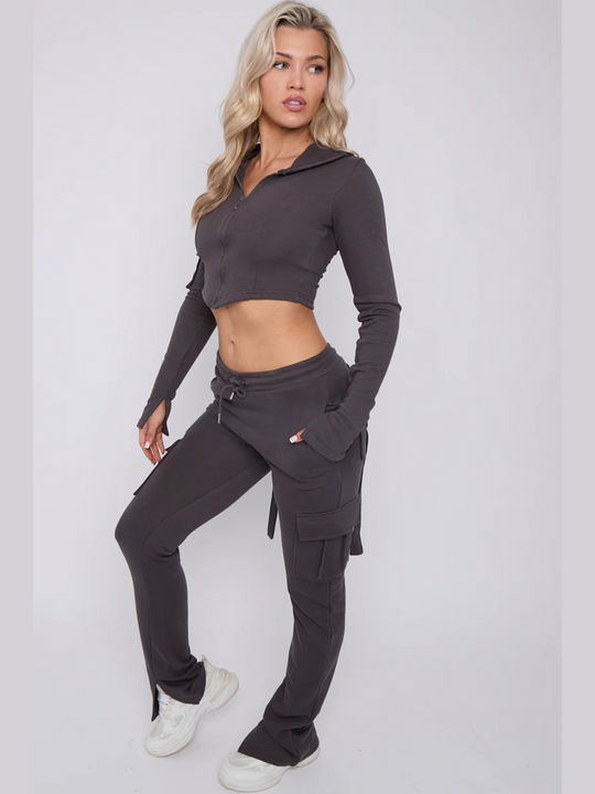 Full length shot Model wears a cropped top zip up  hoodie in  charcoal grey and matching drawstring joggers with a fitted cargo design. Model has blonde hair, she stands to the side with one knee slightly bent, she looks behind her away from the camera. Model wears white trainers. 