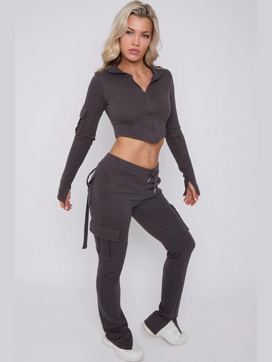 Full length shot. Model wears a cropped top zip up  hoodie in  charcoal grey and matching drawstring joggers with a fitted cargo design. Model has blonde hair,  she stands to the side, the side of the joggers and the loop back design of the back can be seen. Model wears white trainers. 