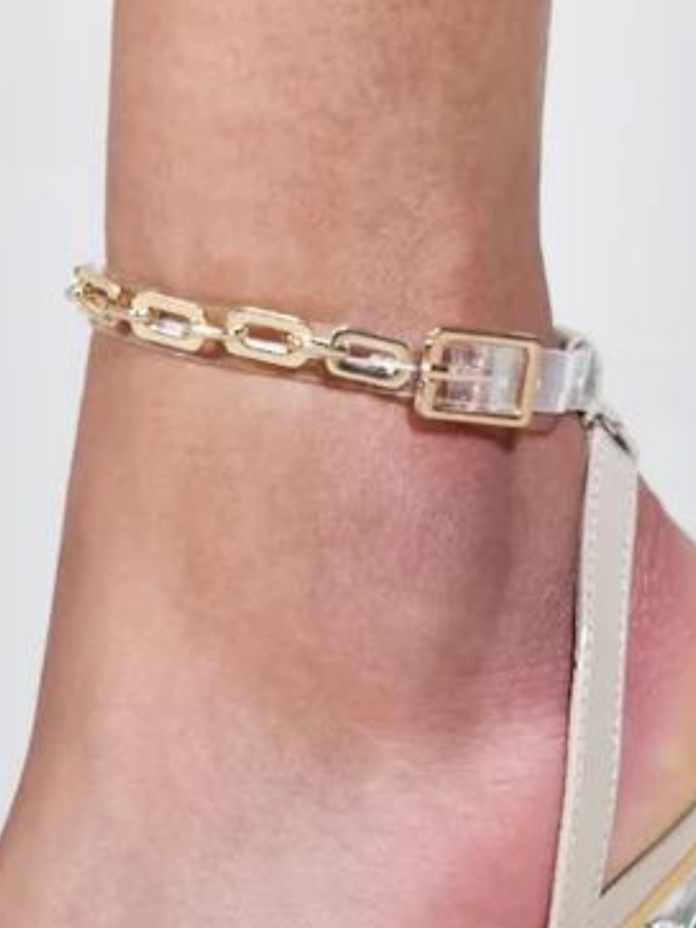 Model wears a beige open toe shoe with a perspex front strap and thin gold link perspex ankle strap, with adjustable strap closure. The beige shoe has a clear stiletto heel. A closeup of the metal chain ankle can be seen.