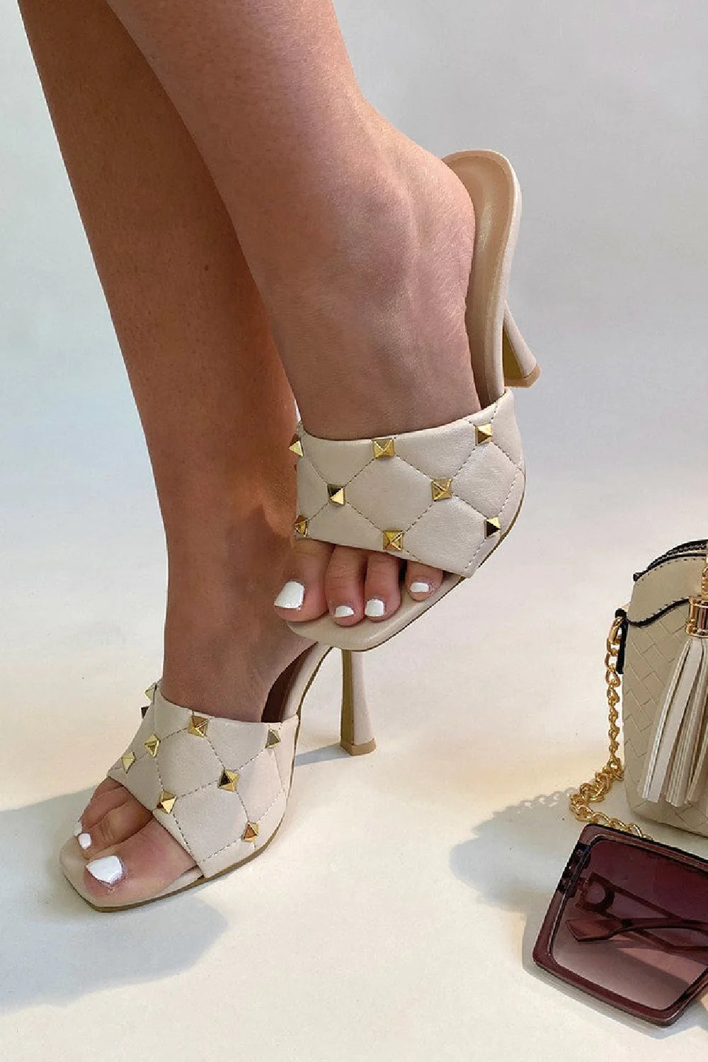 Cream quilted faux leather mule heels with gold studded heels. Model waers cream studded mule heels, she lifts obe leg above the ground. A hand bag and sunglasses are on the ground. 