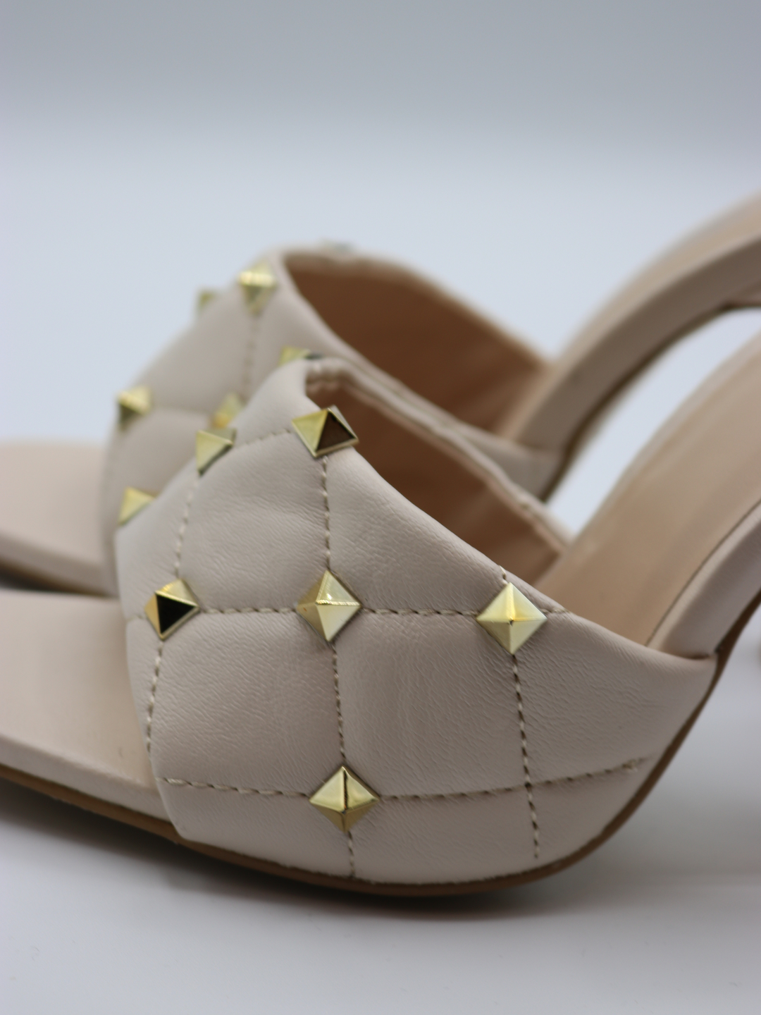 Cream quilted faux leather mule heels with gold studded heels. A closeup of both studded shoes can be seen. 