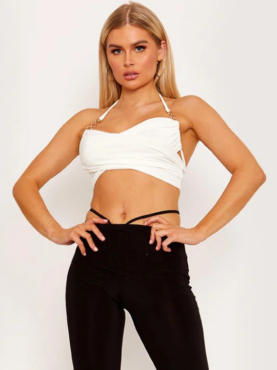 Model wears a white wrap style crop with tie back fastening. The slinky strap has gold chain accents. Model has tanned skin and blonde hair,  she stands slightly to the side, placing both hands on her hips, she looks directly into the camera.