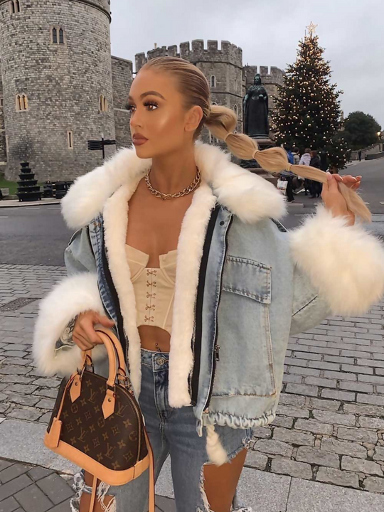Model wears an oversized denim jacket with a white faux fur collar and white faux fur cuffs. Model looks to the side and holds her pony tail in her hand. 