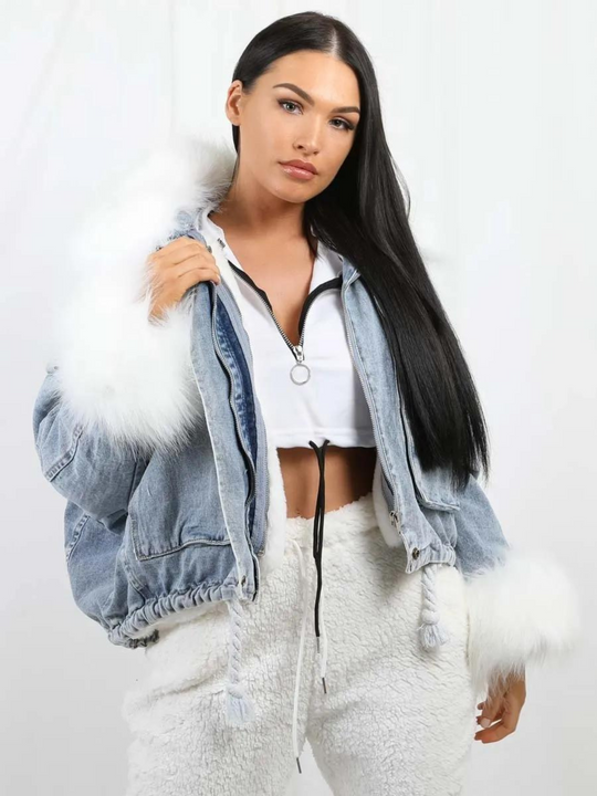 Model wears an oversized denim jacket with a white faux fur collar and white faux fur cuffs. Model wears the jacket open. She wears a white cropped top and white trousers. 