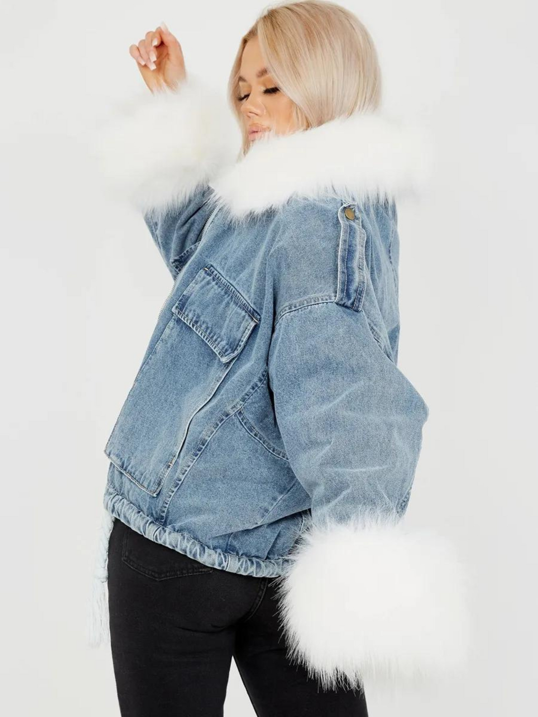 Model wears an oversized denim jacket with a white faux fur collar and white faux fur cuffs. Model has one hand in the air and the other by her side. The white cuffed fur is visible.