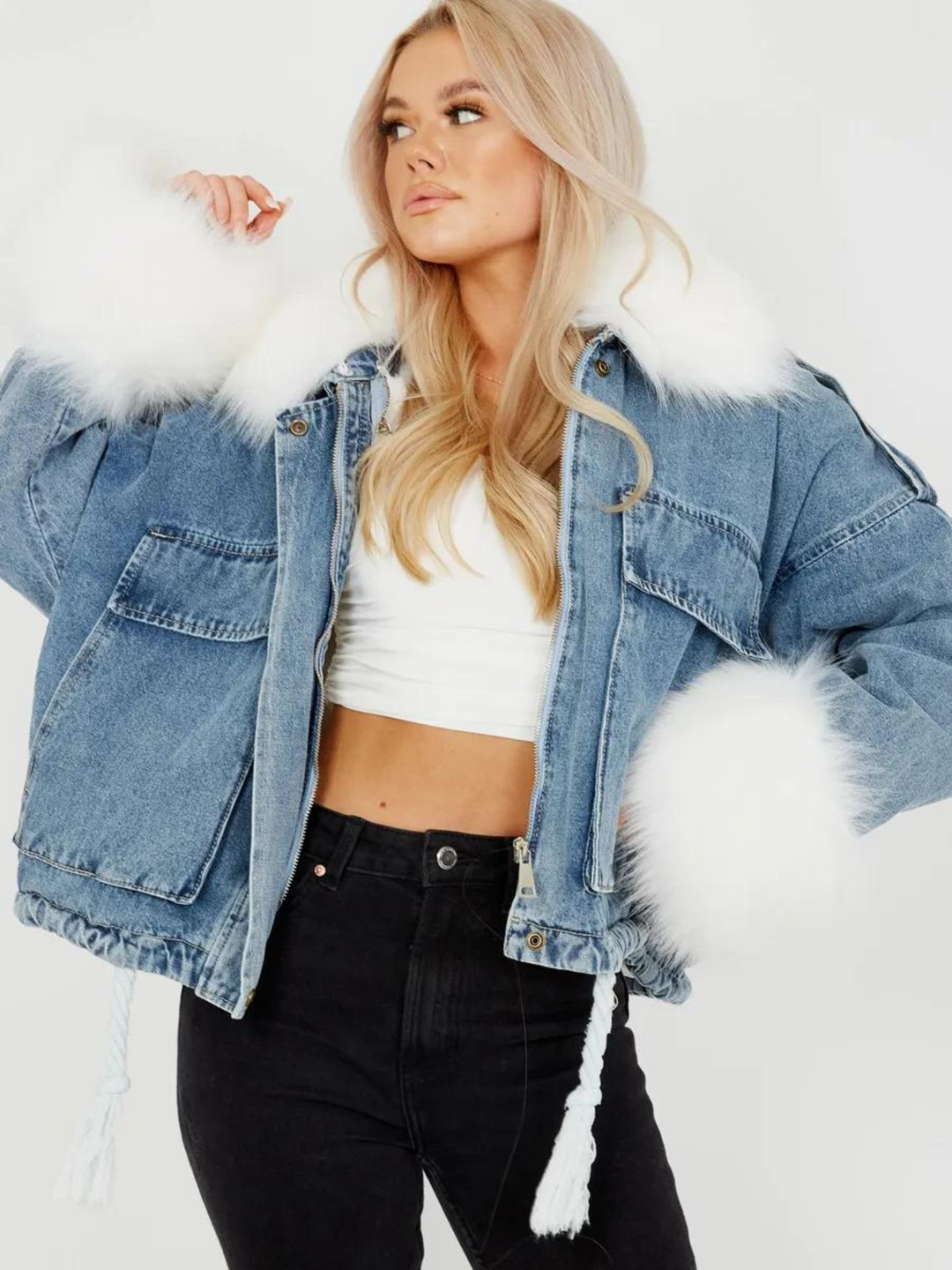 Model wears an oversized denim jacket with a white faux fur collar and white faux fur cuffs. Model has the jacket open and looks to the side. 