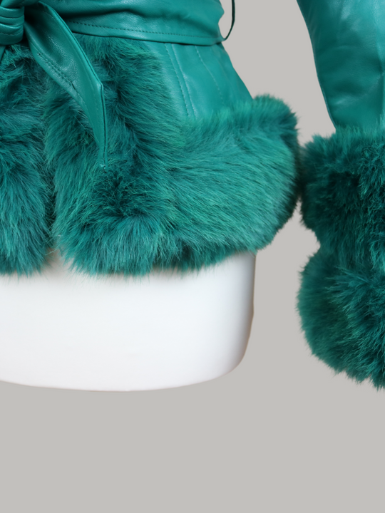 Manequin wears a green faux leather jacket with green faux fur trim. The  green jacket has a faux fur hooded trim and large faux cuffs at the sleeves and a faux leather belt.  The picture shows a close up of the bottom of the jacket including the faur fur trim of the jacket and the faux fur trim at the cuffs of ths sleeves. 