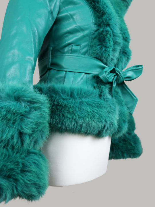 Manequin wears a green faux leather jacket with green faux fur trim. The  green jacket has a faux fur hooded trim and large faux cuffs at the sleeves and a faux leather belt.   The manequin stands to the side, the faux fur cuffes sleeves, faux leather belt and faux fur trim can be seen. 