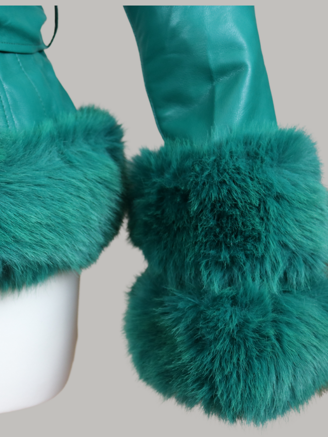 Ghost manequin wears a green faux leather jacket with green faux fur trim. The  green jacket has a faux fur hooded trim and large faux cuffs at the sleeves and a faux leather belt.  The picture shows a close up of the faux fur cuffed sleeves. 