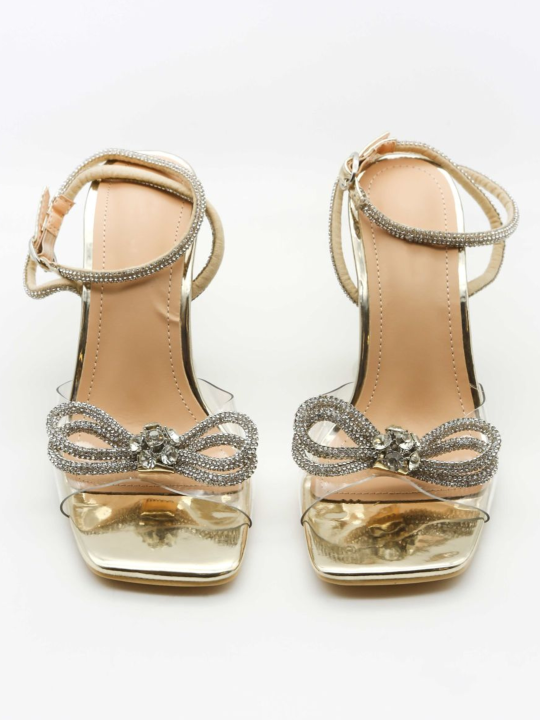Gold open toe heeled shoes, with a front perspex strap, diamante bow front design and diamante ankle strap. The shoes is placed facing forward, the front of the shoes can be seen, including the diamanté bow front and diamanté ankle straps. 