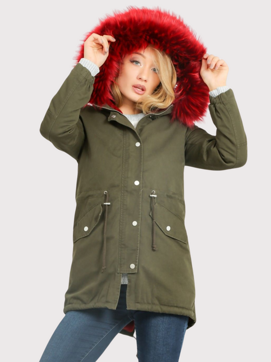Model wears green khaki parka jacket with  burgandy faux fur hooded trim. The coat has a zip and front button fastening and adjustable string tightening. Model wears the faux fur burgandy hood, placing his hands to the hood and looks directly into the camera. 