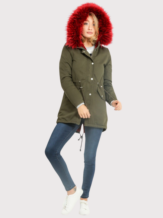 Full length shot. Model wears green khaki parka jacket with  burgandy faux fur hooded trim. The coat has a zip and front button fastening and adjustable string tightening. Model wears the burgandy faux fur hood, she faces the camera, pulling on the adjustable string and bends one leg. Model wears the green parka and blue denim jeans.