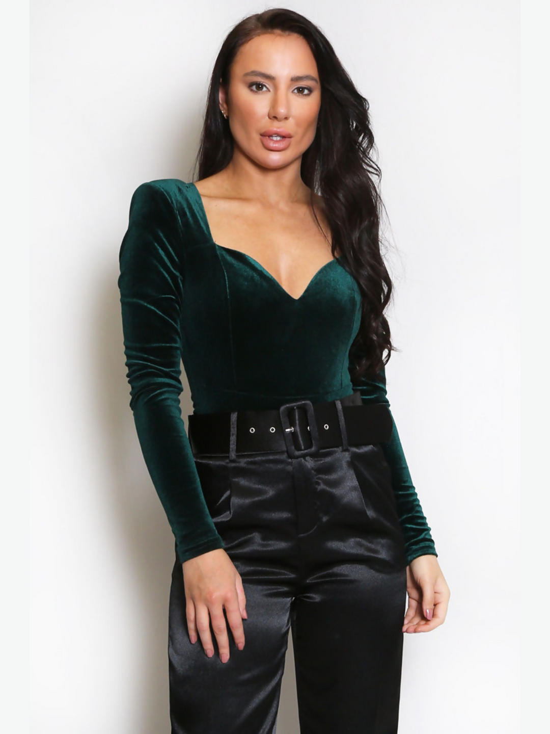 Model wears a luxe green velvet bodysuit with square neck and sweetheart front. The bodysuit has long sleeves and soft padded shoulders. Model also weras a staing belted trousers. Model stands with one hand on her thigh and one hand by her side.