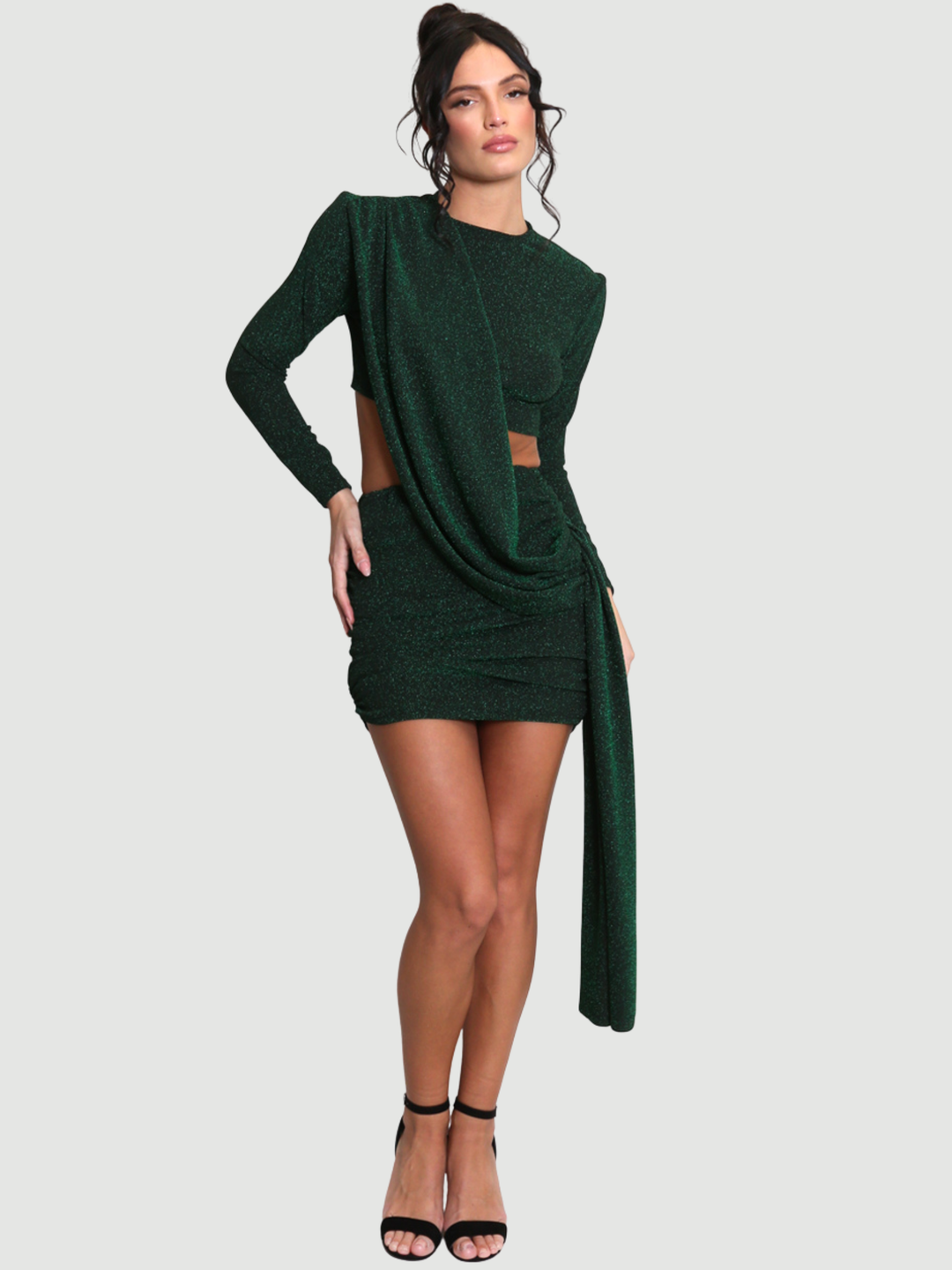 Full length shot. Model stands in front of a plain background. The model wears a cut out lurex green mini dress, with long sleeves and diagnal draped front and rounded neck. The model has tanned skin and black hair that is in a high bun, she faces the camera, placing one hand on her hip and the other by her side, the raped design can be seen. 