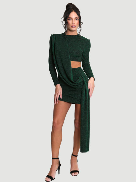 Model stands in front of a plain background. The model wears a cut out lurex green mini dress, with long sleeves and diagnal draped front and rounded neck. The model has tanned skin and black hair that is in a high bun, the draped design can be seen, she places both hands on her thighs.
