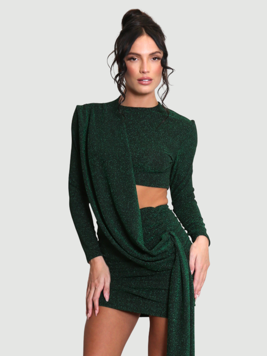 Model stands in front of a plain background. The model wears a cut out lurex green mini dress, with long sleeves and diagnal draped front and rounded neck. The model has tanned skin and black hair that is in a high bun, she faces the camera.