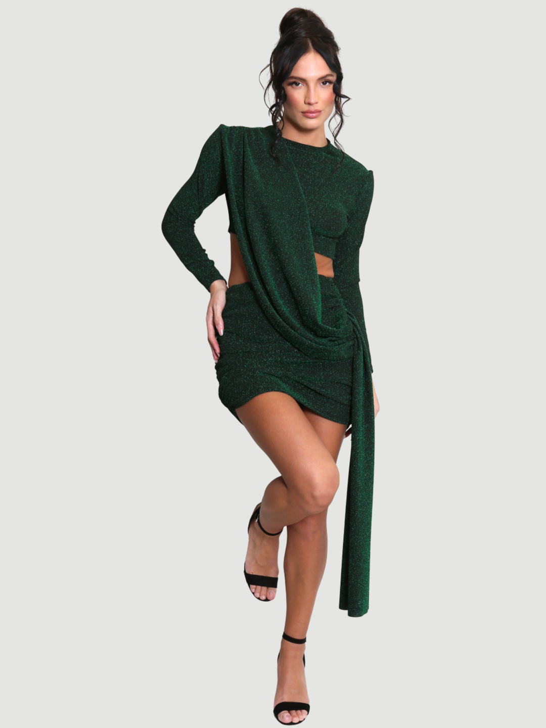 Full length shot. Model stands in front of a plain background. The model wears a cut out lurex green mini dress, with long sleeves and diagnal draped front and rounded neck. The model has tanned skin and black hair that is in a high bun, she places one leg above the gound and the other on her hip, the drape front can be seen.