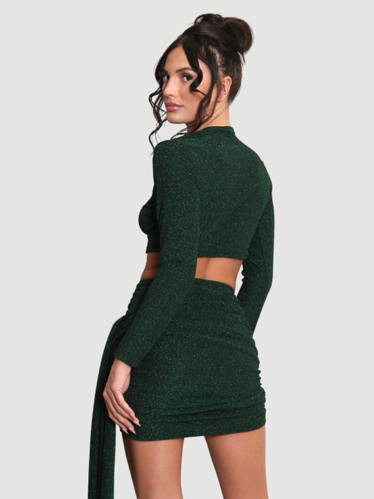 Model stands in front of a plain background. The model wears a cut out lurex green mini dress, with long sleeves and diagnal draped front and rounded neck. The model has tanned skin and black hair that is in a high bun, the model stands with her back to the camera and turns her head to look into the camera. The cut out dress can be seen, including the drape design and lomg sleeves. 