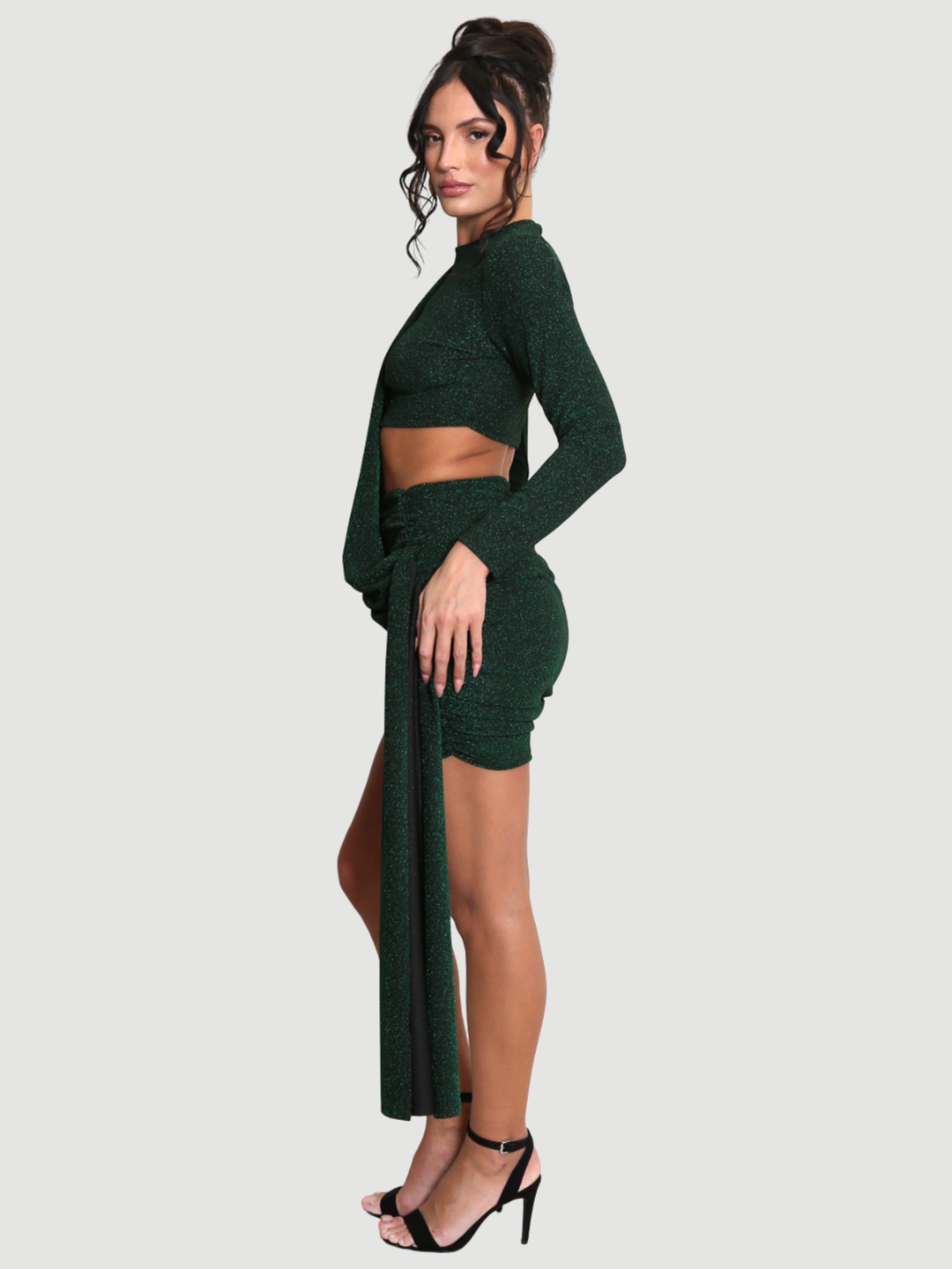Full length shot. Model stands in front of a plain background. The model wears a cut out lurex green mini dress, with long sleeves and diagnal draped front and rounded neck. The model has tanned skin and black hair that is in a high bun. The model stands to the side, the cut out side of the dress can be seen, as well as the draped side.