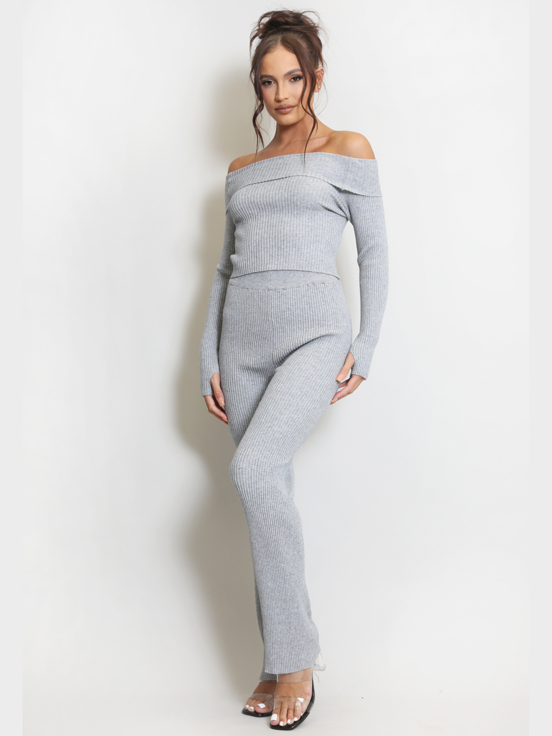Full length shot. Female model stands in front of a plain background. Model wears a grey ribbed long sleeved off the shoulder top, with matching ribbed trousers set.  The model has tanned skin and dark brown hair; she stands with one bent leg and arms by her sides, she wears perspex heels. 