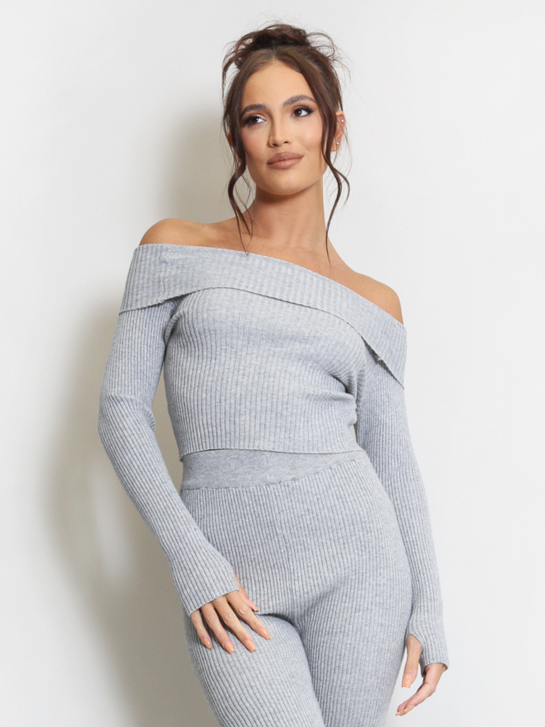 Female model stands in front of a plain background. Model wears a grey ribbed long sleeved off the shoulder top, with matching ribbed trousers set.  The model has tanned skin and dark brown hair, she  faces the camera and places one hand by her sides and the other by her thighs. 