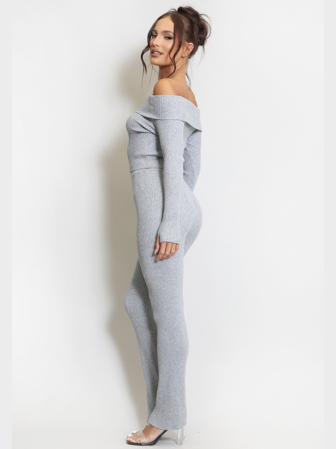 Full length shot. Female model stands in front of a plain background. Model wears a grey ribbed long sleeved off the shoulder top, with matching ribbed trousers set.  The model has tanned skin and dark brown hair, she stands to the side and turns her head to look into the camera. The side of the grey ribbed bardot top and trousers can be seen, she wears perspex heeled shoes at her feet. 