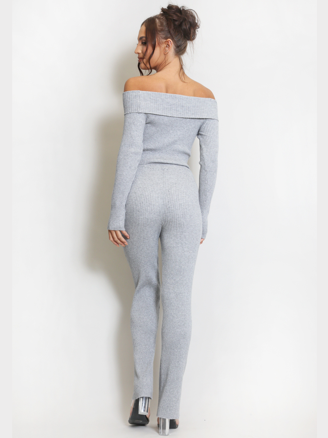 Full length shot. Female model stands in front of a plain background. Model wears a grey ribbed long sleeved off the shoulder top, with matching ribbed trousers set.  The model has tanned skin and dark brown hair, she stands with her back to the camera, the back of the ribbed bardot set can be seen, including the wide legged bottoms. The model wears perspex heeled shoes. 