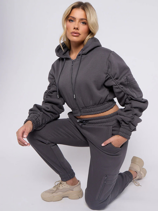 Model wears grey hoodie and matching fitted joggers set. The hoodie has a draw string design, front zip fastening, elasticated waist and ruched sleeves. The matching joggers too has an elasticated waist. The model has blonde hair, she kneels on one knee and places her arm on the other knee, she looks directly into the camera. 