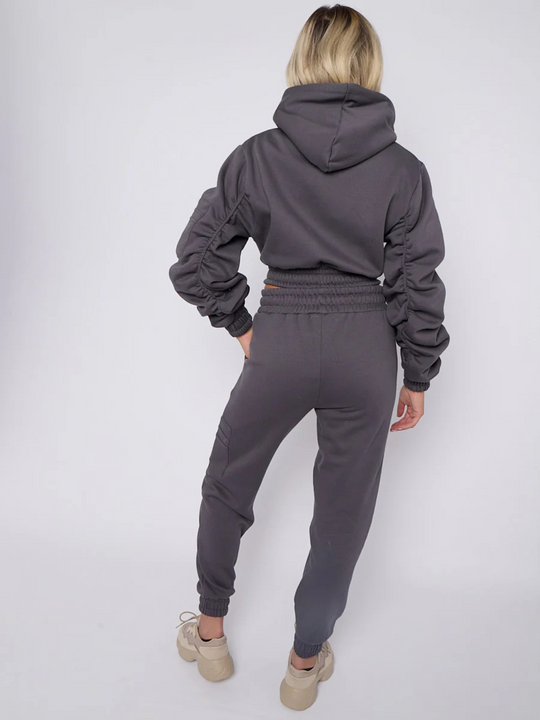 Full length shot. Model wears grey hoodie and matching fitted joggers set. The hoodie has a draw string design, front zip fastening, elasticated waist and ruched sleeves. The matching joggers too has an elasticated waist. The model has blonde hair, she stands with her back to the camera, the hood, ruched sleeves and fitted jumper can be seen. Model wears chunky trainers. 