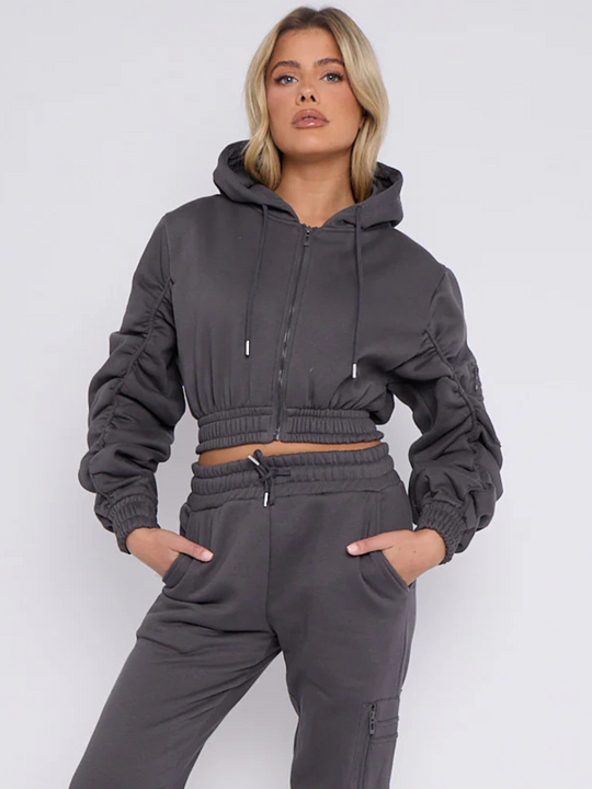 Mid length shot. Model wears grey hoodie and matching fitted joggers set. The hoodie has a draw string design, front zip fastening, elasticated waist and ruched sleeves. The matching joggers too has an elasticated waist. The model has blonde hair,she faces the camera and tilts her head slightly upwards, she places her hands in both jogger pockets and looks directly into the camera, tilting her head upwards slightly. 