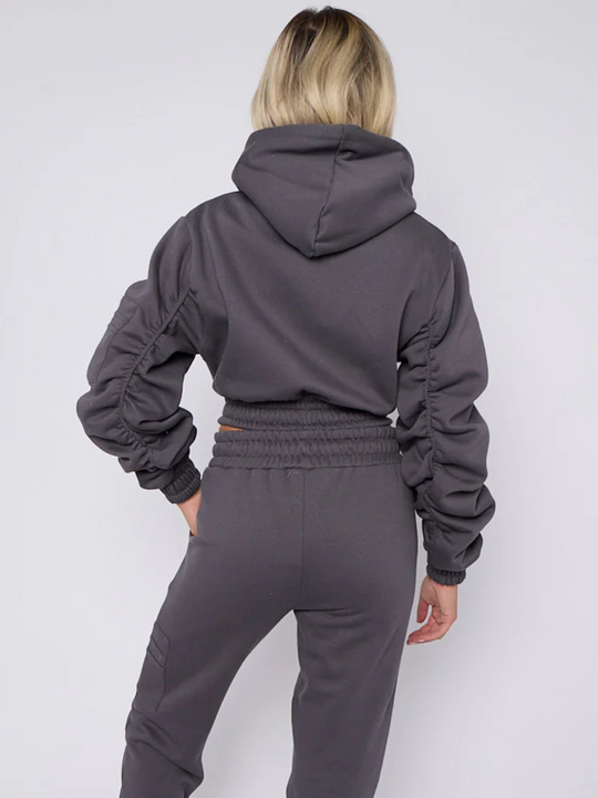 Model wears grey hoodie and matching fitted joggers set. The hoodie has a draw string design, front zip fastening, elasticated waist and ruched sleeves. The matching joggers too has an elasticated waist. The model has blonde hair, she stands with her back to the camera, the ruched sleeves can be seen. 