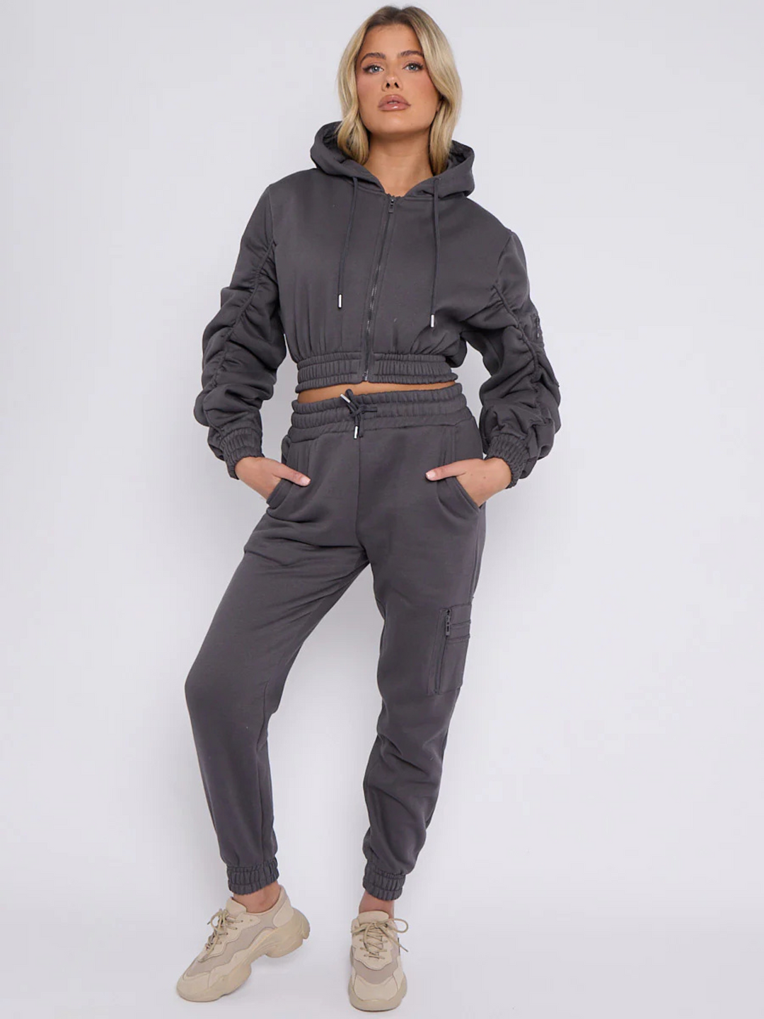 Full length shot. Model wears grey hoodie and matching fitted joggers set. The hoodie has a draw string design, front zip fastening, elasticated waist and ruched sleeves. The matching joggers too has an elasticated waist. The model has blonde hair, she places her ands in her pockets and wears chunky trainers.