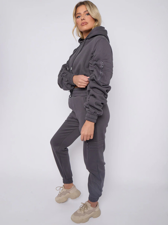 Full length shot. Model wears grey hoodie and matching fitted joggers set. The hoodie has a draw string design, front zip fastening, elasticated waist and ruched sleeves. The matching joggers too has an elasticated waist. The model has blonde hair, she stands to the side, the ruched hoodie sleeves and the side of the joggers can be seen. Model turns her head to look directly into the camera. 