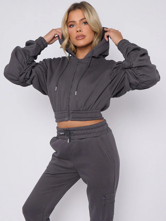 Model wears grey hoodie and matching fitted joggers set. The hoodie has a draw string design, front zip fastening, elasticated waist and ruched sleeves. The matching joggers too has an elasticated waist. The model has blonde hair, she holds her hood on either side of her head and looks directly into the camera. 