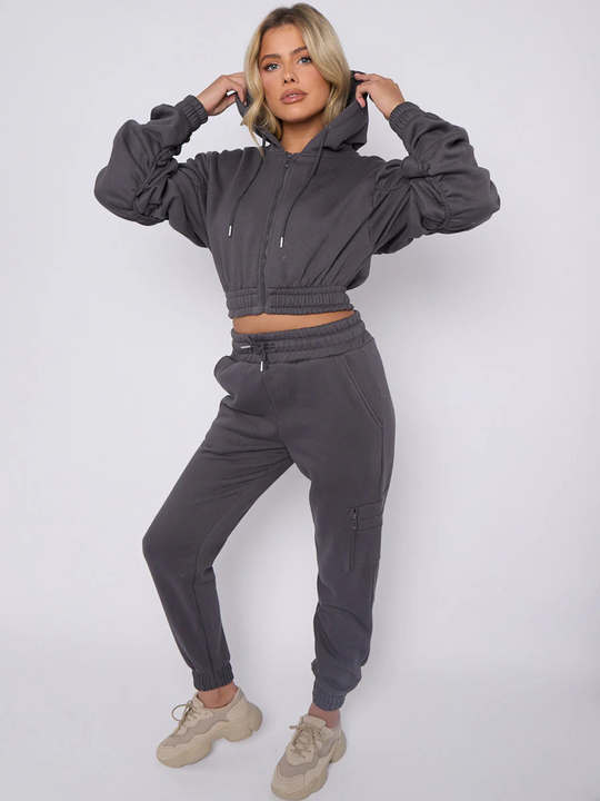 Full length shot. Model wears grey hoodie and matching fitted joggers set. The hoodie has a draw string design, front zip fastening, elasticated waist and ruched sleeves. The matching joggers too has an elasticated waist. The model has blonde hair, she places her hand on either side of the hood. Model stares directly  into the camera. 