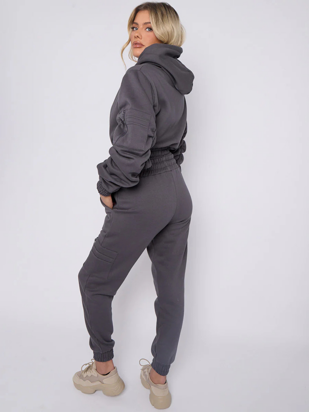 Full length shot. Model wears grey hoodie and matching fitted joggers set. The hoodie has a draw string design, front zip fastening, elasticated waist and ruched sleeves. The matching joggers too has an elasticated waist. The model has blonde hair, she stands to the side and places  her hand in her pocket. Mdel turns her head so that she is looking into the camera, he wears chunky trainers that compliments her jogger set. 