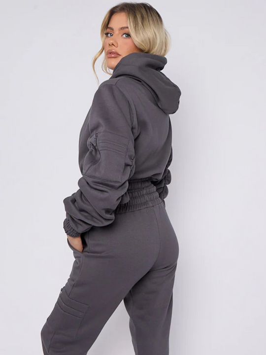 Model wears grey hoodie and matching fitted joggers set. The hoodie has a draw string design, front zip fastening, elasticated waist and ruched sleeves. The matching joggers too has an elasticated waist. The model has blonde hair, model stands to the side and turns her head to face the camera, she places one hand in her pocket, the ruched hooded sleeves can be seen.