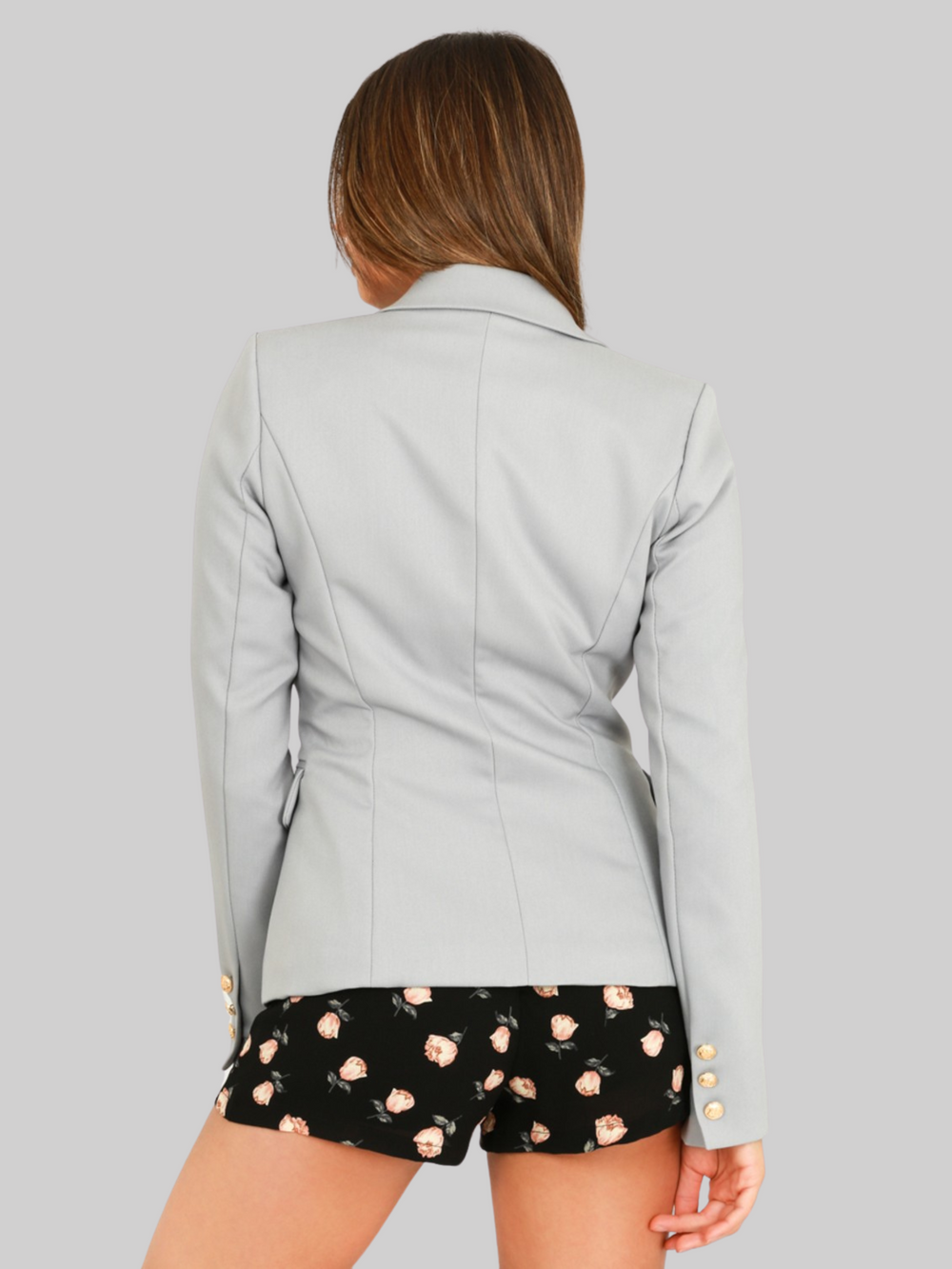 female model wears a grey double breatsted blazer with gold military buttons at the front of the blazer and the cuffs. The model has tanned skin and long brown hair, the model stands with her bak to the camera, the back of the blazer and the miltary cuff detailing can be seen. 
