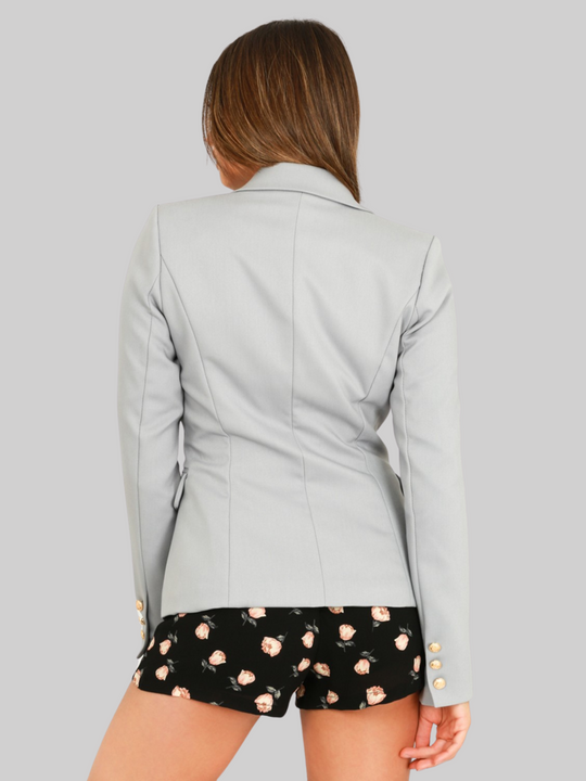 female model wears a grey double breatsted blazer with gold military buttons at the front of the blazer and the cuffs. The model has tanned skin and long brown hair, the model stands with her bak to the camera, the back of the blazer and the miltary cuff detailing can be seen. 