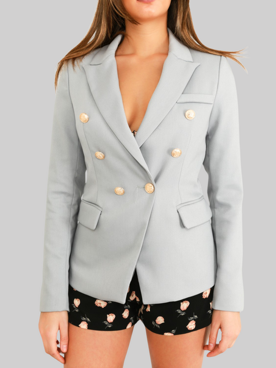 Torso shot. Female model wears a grey double breatsted blazer with gold military buttons at the front of the blazer and the cuffs. The model has tanned skin and long brown hair, the blazer is closed with the military button fastening, she stands with her hands to her side. 