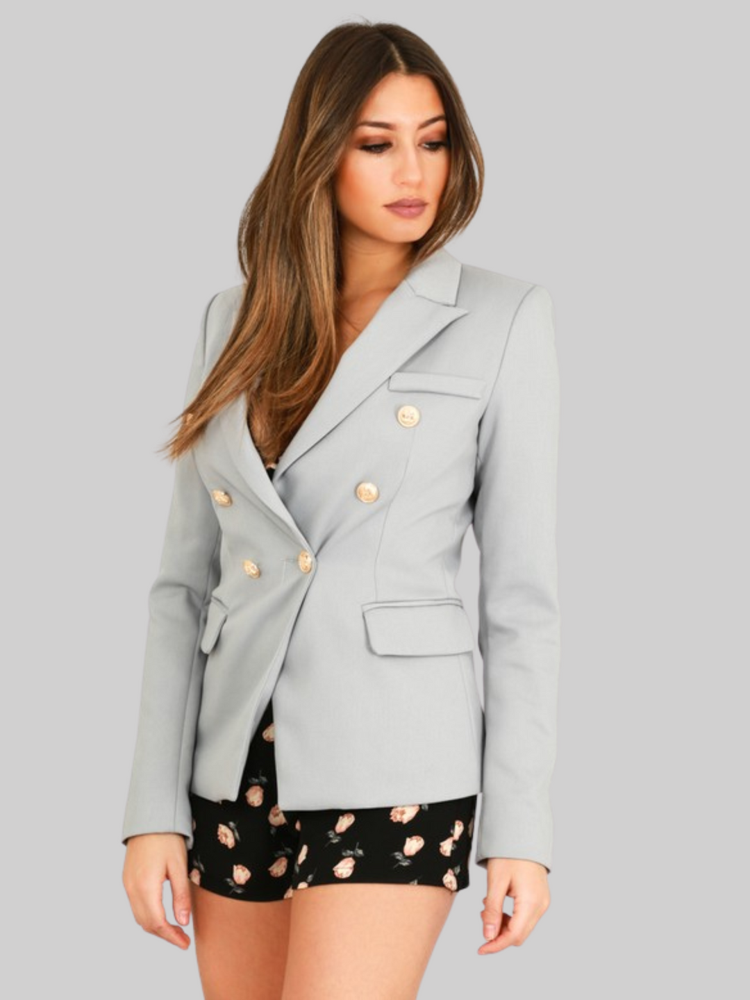 Female model wears a grey double breatsted blazer with gold military buttons at the front of the blazer and the cuffs. The model has tanned skin and long brown hair, she looks away from the camera and wears the military diuvle breasted blazer closed. 
